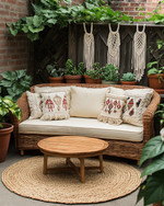 A cozy boho patio featuring a low-profile rattan loveseat with cream cushions, layered with macrame and embroidered pillows. A round jute rug sits beneath a small wooden coffee table, surrounded by various potted plants in terracotta and ceramic planters, including a trailing Pothos in a macrame hanger. — alternate angle