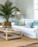 A bright coastal poolhouse interior with a white slipcovered sofa, rattan armchair, and light wood coffee table, accented with soft blue and green pillows, a jute rug, and a large potted palm plant. — close-up of textures and materials