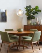 A cozy Mid-Century Modern dining room featuring a round walnut dining table with four upholstered chairs in a muted olive green boucle fabric. A George Nelson bubble lamp hangs above, casting a soft glow. A vintage teak credenza against the wall displays ceramic vases and a large Fiddle Leaf Fig plant. — alternate angle
