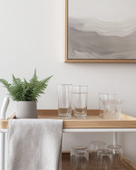 A serene Scandinavian home bar setup in a rental apartment featuring a light wood bar cart with white metal accents, minimalist glassware, and a small potted fern. Soft grey wall art and a textured linen tea towel complete the inviting, renter-friendly space. — close-up of textures and materials