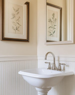 A traditional powder room featuring a white pedestal sink, polished nickel faucet, and beadboard wainscoting painted in a warm off-white. A large framed mirror hangs above, reflecting a botanical print on the opposite wall, all under soft, warm lighting. — styling and décor detail