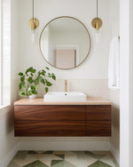 A Mid-Century Modern bathroom featuring a walnut floating vanity with an integrated white sink, brushed brass fixtures, and a large round mirror. The walls are painted a soft off-white, complemented by geometric floor tiles in muted green and cream. A trailing Pothos plant adds a touch of organic warmth. — close-up of textures and materials