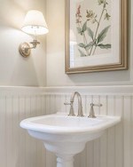 A traditional powder room featuring a white pedestal sink, polished nickel faucet, and beadboard wainscoting painted in a warm off-white. A large framed mirror hangs above, reflecting a botanical print on the opposite wall, all under soft, warm lighting. — close-up of textures and materials