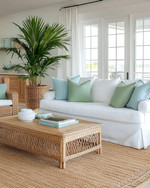 A bright coastal poolhouse interior with a white slipcovered sofa, rattan armchair, and light wood coffee table, accented with soft blue and green pillows, a jute rug, and a large potted palm plant. — styling and décor detail