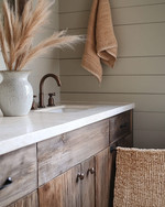A cozy rustic powder room featuring a reclaimed wood vanity with an off-white quartz countertop, an oil-rubbed bronze faucet, and shiplap walls painted in a soft greige. A woven jute rug sits on the floor, and a ceramic vase with dried pampas grass adorns the counter. — styling and décor detail