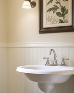 A traditional powder room featuring a white pedestal sink, polished nickel faucet, and beadboard wainscoting painted in a warm off-white. A large framed mirror hangs above, reflecting a botanical print on the opposite wall, all under soft, warm lighting. — alternate angle