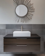 A modern powder room features a floating dark wood vanity with an integrated white quartz sink and brushed nickel faucet, set against large format charcoal grey porcelain tiles. A simple round mirror and abstract metal wall art complete the sleek, resilient design.
