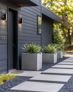 A modern exterior featuring a fresh coat of deep charcoal grey paint on the siding, a contrasting matte black front door, and sleek brushed nickel house numbers. The pathway combines light grey concrete pavers with dark pea gravel, and large concrete planters flank the entrance with sculptural green plants. — alternate angle
