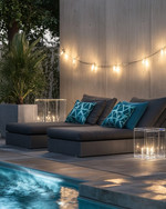 Modern poolside area with low-profile charcoal grey modular loungers, clear acrylic side tables, and large faux concrete planters. The scene is illuminated by sleek rechargeable LED lanterns and subtle solar-powered string lights, with deep teal and slate blue geometric outdoor cushions adding pops of color. — styling and décor detail