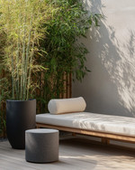 A small Japandi backyard featuring a low-profile teak bench with off-white cushions, a round grey stone side table, and a tall bamboo plant in a black ceramic planter, all set on light wood decking. — styling and décor detail