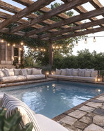 A cozy farmhouse pool area featuring a large swimming pool with natural stone coping, surrounded by reclaimed wood pergola. Plush outdoor sofas with cream cushions and striped pillows are arranged on a flagstone patio, illuminated by Edison bulb string lights overhead and decorative lanterns. — alternate angle