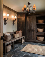 A rustic mudroom featuring warm layered lighting from a wrought iron chandelier and wall sconces, with slate floor tiles, a reclaimed wood bench, and dark wood storage cabinets, accented by woven baskets and a jute rug. — alternate angle