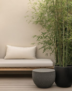 A small Japandi backyard featuring a low-profile teak bench with off-white cushions, a round grey stone side table, and a tall bamboo plant in a black ceramic planter, all set on light wood decking. — alternate angle