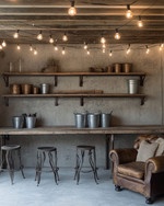 A rustic garage interior featuring exposed concrete walls, a salvaged wood workbench with metal stools, overhead string lights with Edison bulbs, and open shelving made from iron pipes and rough-sawn lumber, accented with galvanized buckets and a worn leather armchair.