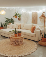 A cozy boho basement living area with a light cream sectional sofa, a round jute rug, and a natural wood coffee table. Warm lighting from a rattan floor lamp and string lights illuminates various potted plants, macrame wall hangings, and terracotta accents. — alternate angle