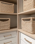 A clean coastal minimalist closet featuring light oak shelving, white built-in drawers with brushed nickel pulls, and tightly woven seagrass baskets, all bathed in natural light. — styling and décor detail