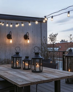 Industrial deck at dusk featuring warm white Edison bulb string lights draped overhead, dark bronze gooseneck wall sconces, a raw wood dining table, and large black metal lanterns with LED candles. — close-up of textures and materials