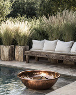 A rustic poolside area featuring a reclaimed timber bench with linen cushions, large natural stone planters filled with tall grasses, and a hammered copper fire bowl, all set against a backdrop of natural stone paving and lush greenery.