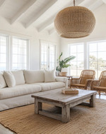 A bright coastal great room featuring a large off-white linen sofa, a reclaimed wood coffee table, and a natural jute area rug. Rattan armchairs flank the space, while a large woven pendant light hangs from the ceiling, all under ample natural light.
