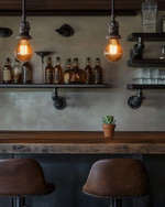 An inviting industrial home bar featuring a reclaimed wood countertop, dark metal pipe shelving, and leather bar stools. Warm Edison bulb pendants cast an amber glow, highlighting vintage amber glass bottles and a small potted succulent. — styling and décor detail