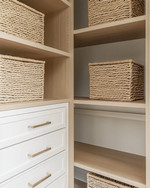A clean coastal minimalist closet featuring light oak shelving, white built-in drawers with brushed nickel pulls, and tightly woven seagrass baskets, all bathed in natural light. — alternate angle