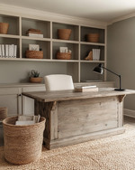 A warm farmhouse home office featuring a large distressed oak desk, a cream linen office chair, open shelving with books and woven baskets, and walls painted in a soft greige. A braided jute rug grounds the space, and a matte black task lamp sits on the desk. — close-up of textures and materials
