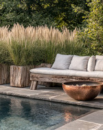 A rustic poolside area featuring a reclaimed timber bench with linen cushions, large natural stone planters filled with tall grasses, and a hammered copper fire bowl, all set against a backdrop of natural stone paving and lush greenery. — styling and décor detail