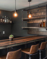 An inviting industrial home bar featuring a reclaimed wood countertop, dark metal pipe shelving, and leather bar stools. Warm Edison bulb pendants cast an amber glow, highlighting vintage amber glass bottles and a small potted succulent. — alternate angle