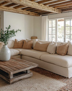 A cozy farmhouse poolhouse interior featuring a large cream sectional sofa with woven throw pillows, a distressed wood coffee table, and an exposed reclaimed wood ceiling. Soft light filters in, highlighting a natural fiber rug and a simple ceramic vase with greenery.
