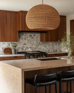 A Mid-Century Modern kitchen featuring flat-panel walnut cabinetry, a honed quartzite island, and a geometric terrazzo backsplash. A large woven rattan pendant light hangs above the island, which is flanked by black leather bar stools, all in soft natural daylight.