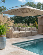 A serene coastal pool area featuring light travertine pavers, deep-seated wicker outdoor sofas with sand-colored cushions, and a large cantilever umbrella providing ample shade. Lush green ornamental grasses in weighty ceramic planters frame the space, while a weathered wood outdoor shower stands nearby. — styling and décor detail