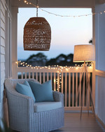 A well-lit coastal porch at dusk, featuring a woven rattan pendant light, string lights draped along the railing, and a floor lamp with a linen shade next to a white wicker chair with soft blue throw pillows. — styling and décor detail