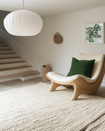 A bright Scandinavian basement living area featuring a light oak sculptural lounge chair, a large cream hand-knotted wool rug, and an oversized white paper lantern pendant light, with subtle forest green textile accents. — styling and décor detail