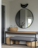 A modern mudroom features a natural oak console table with a minimalist black metal frame, paired with a charcoal grey upholstered bench. Overhead, a matte black sculptural flush-mount light illuminates the space, complemented by a large, frameless circular mirror above the console and woven jute baskets for storage. — alternate angle