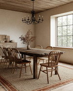 A rustic dining room featuring a large, solid reclaimed oak dining table, mismatched wooden chairs, and a natural jute area rug, illuminated by a wrought iron chandelier, with walls painted in a warm antique white. — styling and décor detail