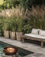 A rustic poolside area featuring a reclaimed timber bench with linen cushions, large natural stone planters filled with tall grasses, and a hammered copper fire bowl, all set against a backdrop of natural stone paving and lush greenery. — alternate angle