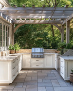 A refined traditional outdoor kitchen featuring off-white Shaker-style cabinetry, honed Pietra Cardosa countertops, and integrated stainless steel appliances under a simple pergola, with natural stone pavers.