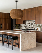 A Mid-Century Modern kitchen featuring flat-panel walnut cabinetry, a honed quartzite island, and a geometric terrazzo backsplash. A large woven rattan pendant light hangs above the island, which is flanked by black leather bar stools, all in soft natural daylight. — close-up of textures and materials