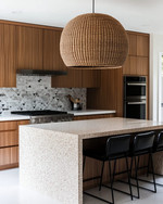 A Mid-Century Modern kitchen featuring flat-panel walnut cabinetry, a honed quartzite island, and a geometric terrazzo backsplash. A large woven rattan pendant light hangs above the island, which is flanked by black leather bar stools, all in soft natural daylight. — styling and décor detail