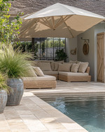 A serene coastal pool area featuring light travertine pavers, deep-seated wicker outdoor sofas with sand-colored cushions, and a large cantilever umbrella providing ample shade. Lush green ornamental grasses in weighty ceramic planters frame the space, while a weathered wood outdoor shower stands nearby. — alternate angle