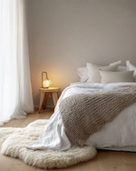 A cozy Scandinavian bedroom featuring a bed dressed in white linen, a chunky knit throw, and a sheepskin rug on light wooden floorboards. Soft, diffused light comes from a simple bedside lamp and a large window. — styling and décor detail