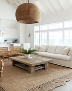 A bright coastal great room featuring a large off-white linen sofa, a reclaimed wood coffee table, and a natural jute area rug. Rattan armchairs flank the space, while a large woven pendant light hangs from the ceiling, all under ample natural light. — close-up of textures and materials