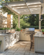 A refined traditional outdoor kitchen featuring off-white Shaker-style cabinetry, honed Pietra Cardosa countertops, and integrated stainless steel appliances under a simple pergola, with natural stone pavers. — close-up of textures and materials