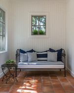A compact traditional poolhouse interior featuring white beadboard walls, a small daybed with navy and white striped cushions, a wrought iron side table, and terracotta tile flooring, bathed in natural light. — alternate angle