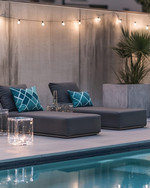 Modern poolside area with low-profile charcoal grey modular loungers, clear acrylic side tables, and large faux concrete planters. The scene is illuminated by sleek rechargeable LED lanterns and subtle solar-powered string lights, with deep teal and slate blue geometric outdoor cushions adding pops of color. — alternate angle