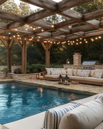 A cozy farmhouse pool area featuring a large swimming pool with natural stone coping, surrounded by reclaimed wood pergola. Plush outdoor sofas with cream cushions and striped pillows are arranged on a flagstone patio, illuminated by Edison bulb string lights overhead and decorative lanterns.