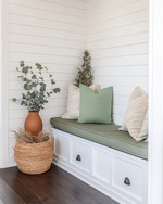 A bright farmhouse mudroom with white shiplap walls, a built-in bench with a sage green cushion, and dark wood flooring. Seasonal touches include a vase of eucalyptus, woven baskets, and a terracotta pot with dried botanicals, all bathed in warm, natural light. — styling and décor detail