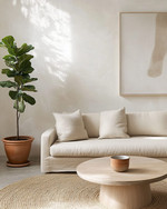 A serene Japandi great room featuring a low-slung oatmeal linen sofa, a light oak round coffee table, and a natural jute rug. Walls are painted in a soft off-white, adorned with a minimalist abstract nature print. A tall Fiddle Leaf Fig in a terracotta pot stands beside a ceramic table lamp with a linen shade, creating a calm and inviting atmosphere. — styling and décor detail