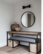 A modern mudroom features a natural oak console table with a minimalist black metal frame, paired with a charcoal grey upholstered bench. Overhead, a matte black sculptural flush-mount light illuminates the space, complemented by a large, frameless circular mirror above the console and woven jute baskets for storage. — close-up of textures and materials