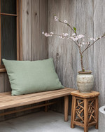 A Japandi porch blending vintage and modern, featuring a low-slung teak bench with a sage green linen cushion, a small hand-carved wooden stool as a side table, and a vintage Japanese stoneware vase with a single cherry blossom branch, all set against a backdrop of weathered wood and concrete planters. — close-up of textures and materials
